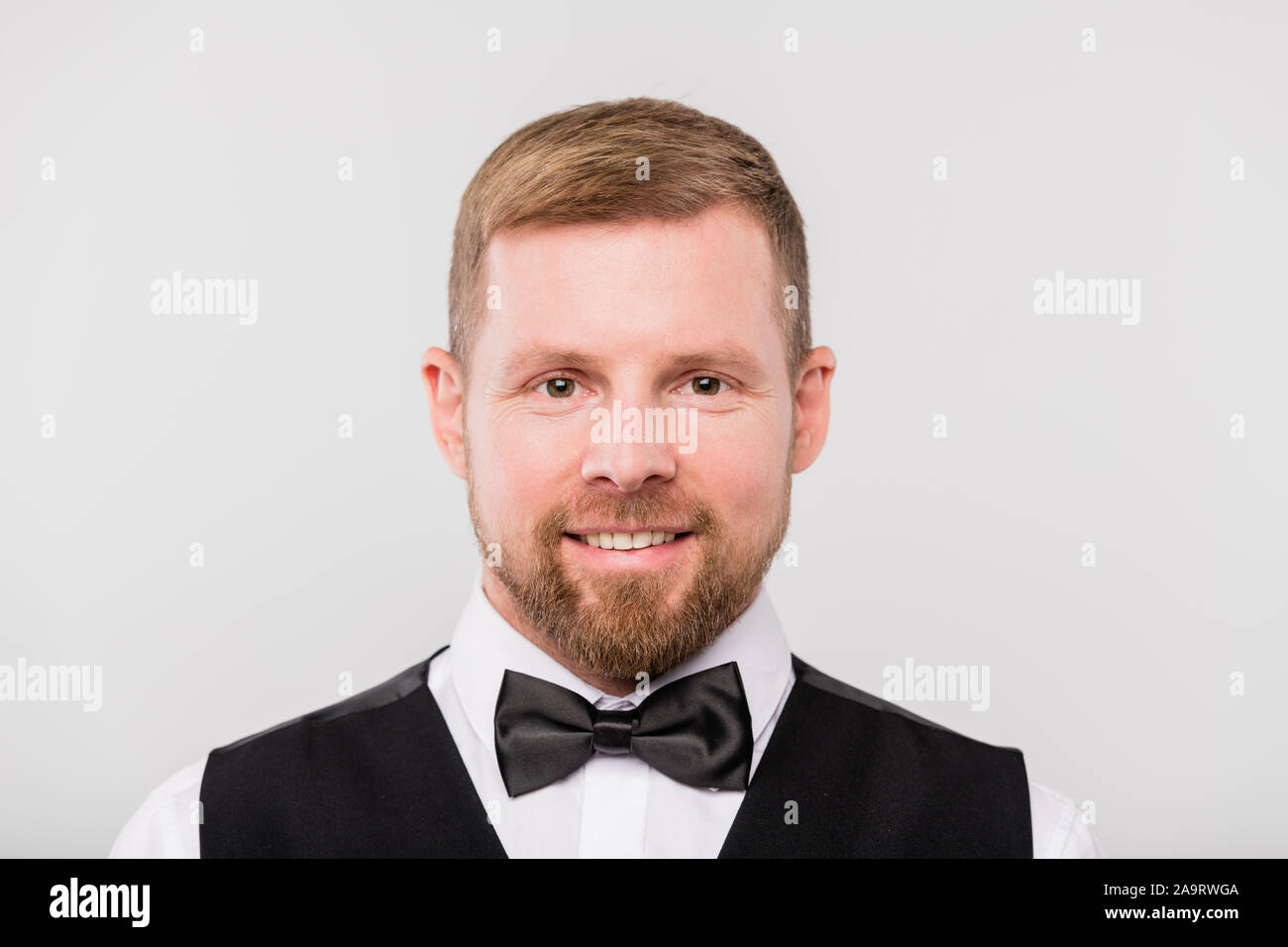 Young smiling bearded waiter in black waistcoat and bowtie looking at you Stock Photo