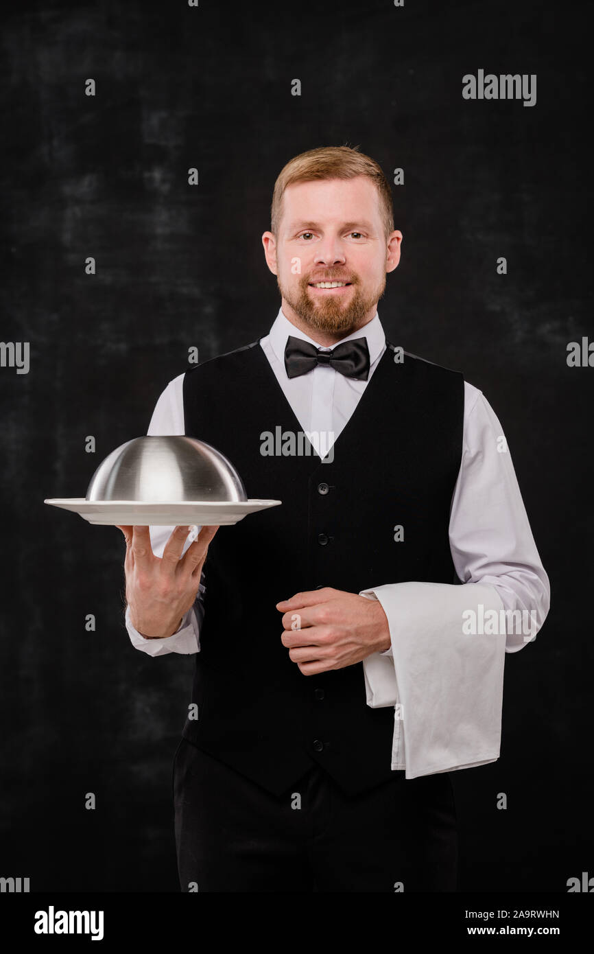 Happy young elegant waiter holding white towel and cloche with food Stock Photo