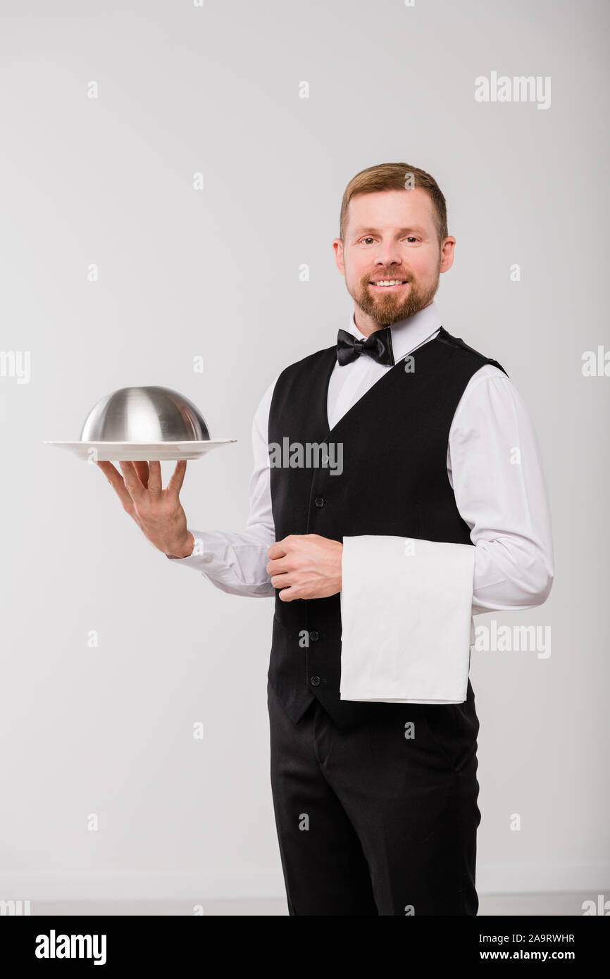 Happy young waiter in black waistcoat and bowtie holding white towel and cloche Stock Photo