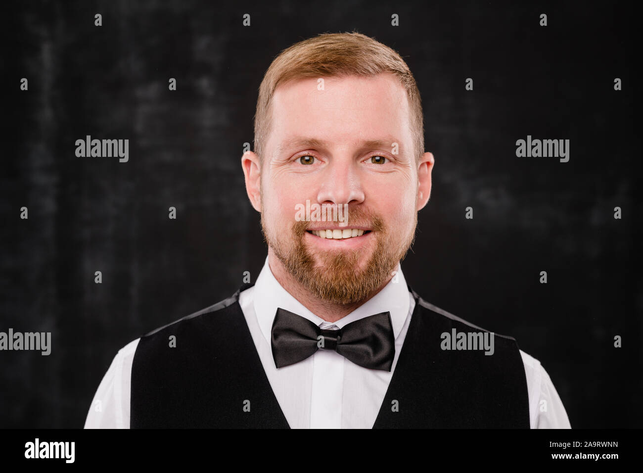 Happy young elegant waiter in black waistcoat and bowtie looking at you Stock Photo