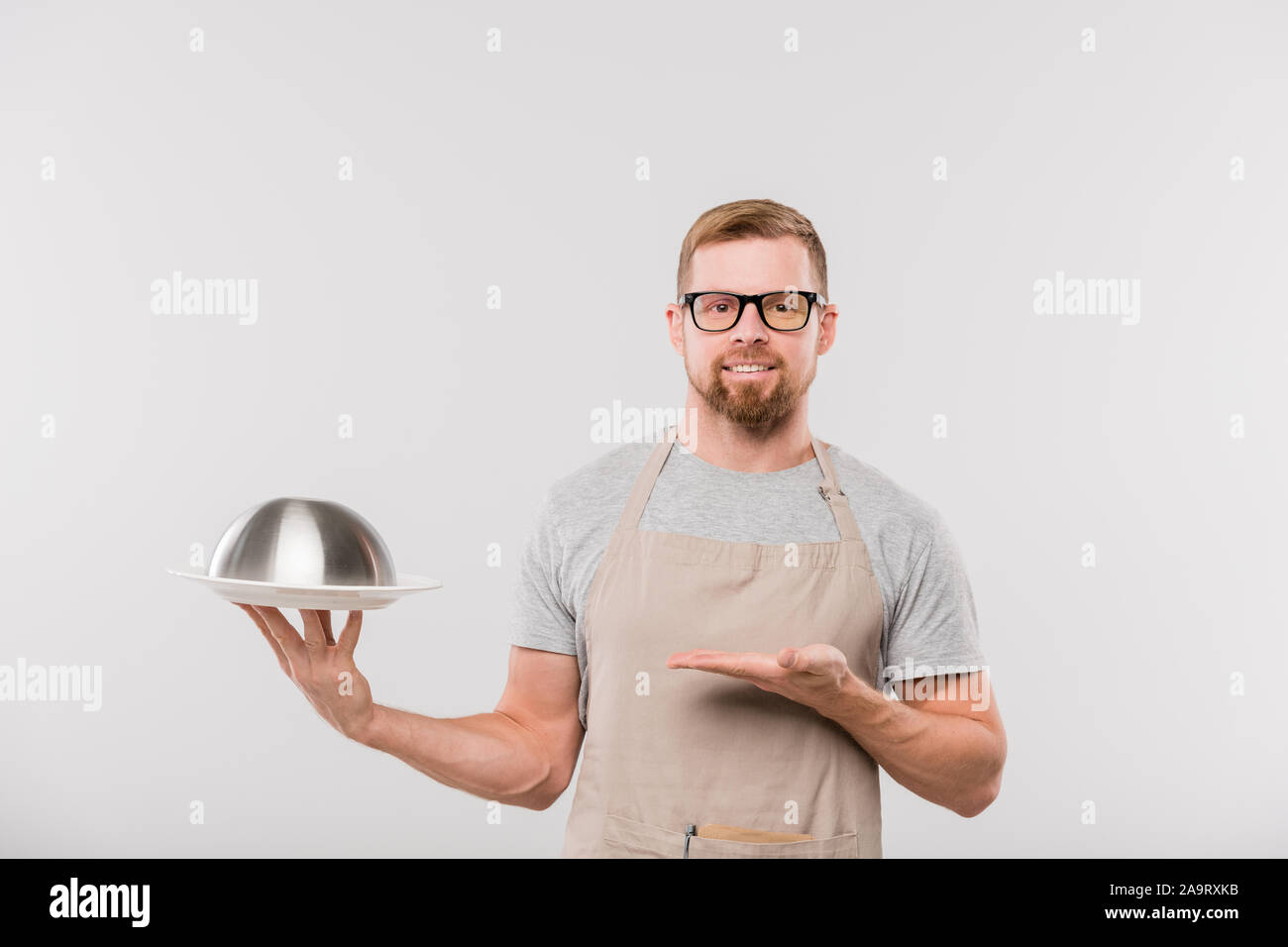 Happy young bearded waiter in apron holding cloche with cooked food Stock Photo