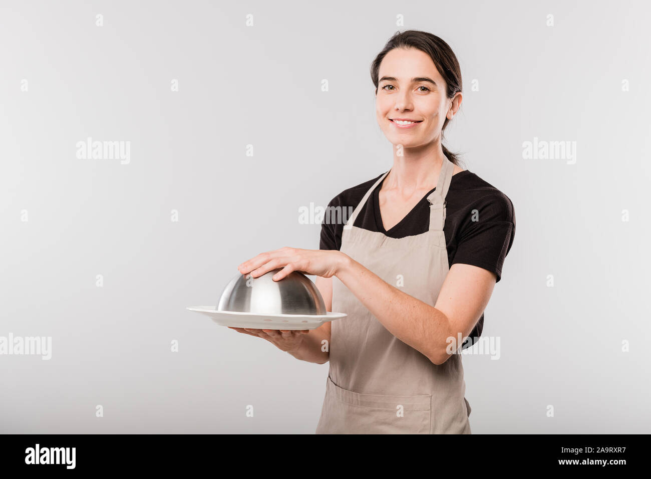 Happy young brunette waitress in apron keeping hand on cover of cloche Stock Photo