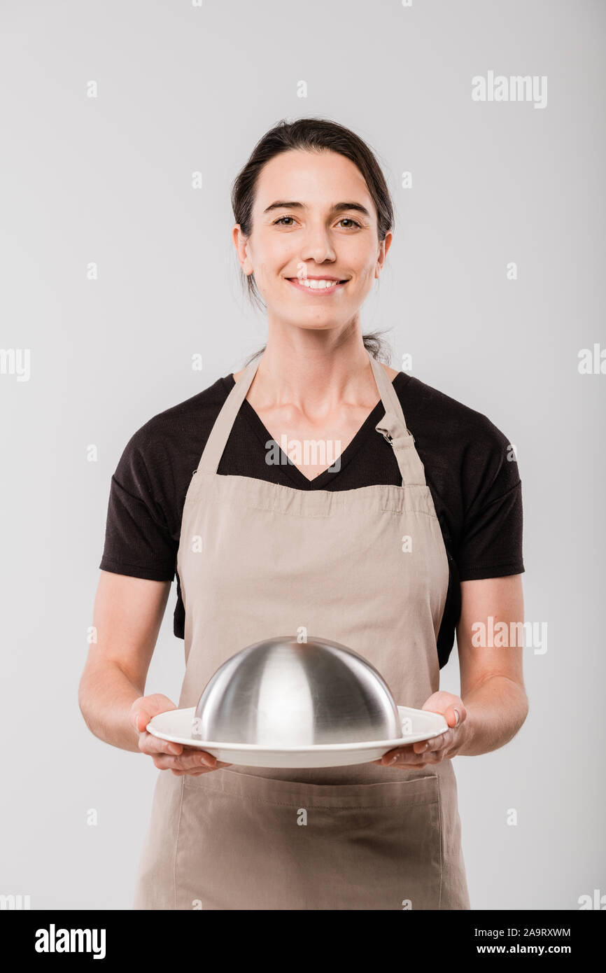 Happy young brunette waitress holding cloche with prepared food for client Stock Photo