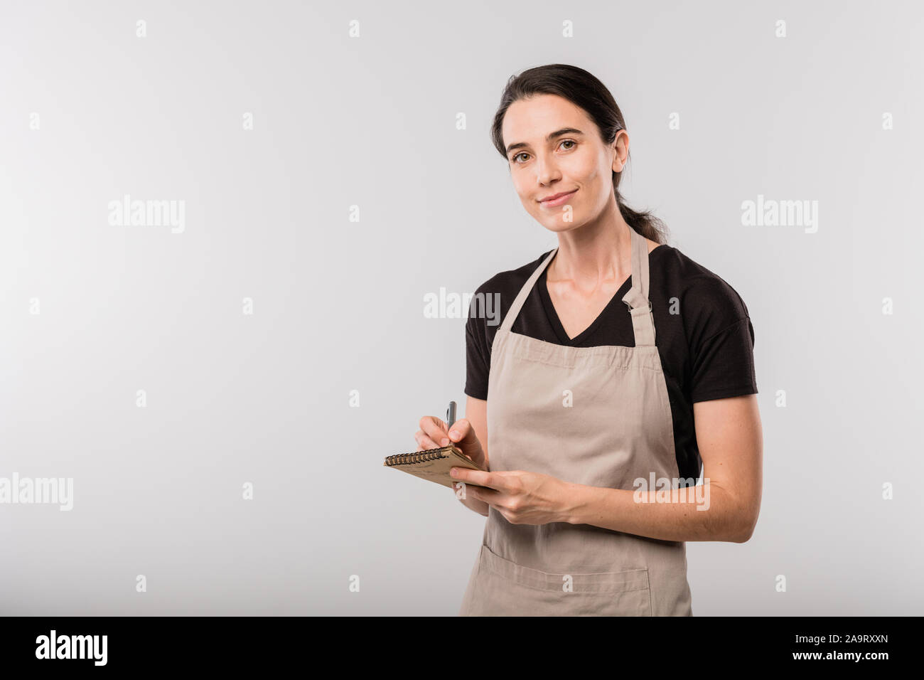 Pretty young brunette waitress in apron making notes about order in notepad Stock Photo
