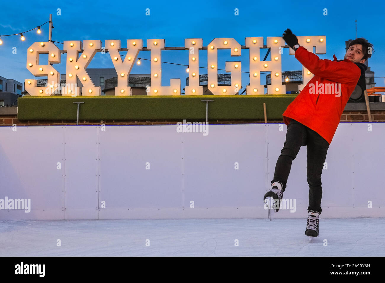 Tobacco Dock, London, UK, 17th November 2019. Skylight staffer Peter goes for a spin on the ice, and a little 'joke fall' in front of the illuminated 'Skylight' sign. People enjoy spectacular views across to the City of London at London's trendy Skylight ice rink, a rooftop at the historic Tobacco Dock complex in Wapping, East London. Credit: Imageplotter/Alamy Live News Stock Photo