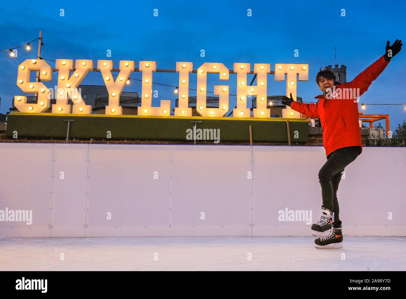 Tobacco Dock, London, UK, 17th November 2019. Skylight staffer Peter goes for a spin on the ice in front of the illuminated 'Skylight' sign. People enjoy spectacular views across to the City of London at London's trendy Skylight ice rink, a rooftop at the historic Tobacco Dock complex in Wapping, East London. Credit: Imageplotter/Alamy Live News Stock Photo