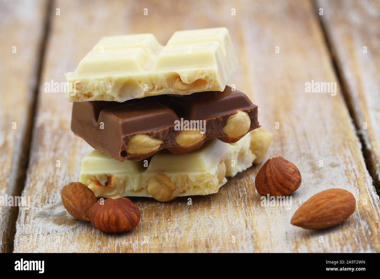 Pieces of white and milk chocolates with almonds and hazelnuts stacked up on wooden surface Stock Photo