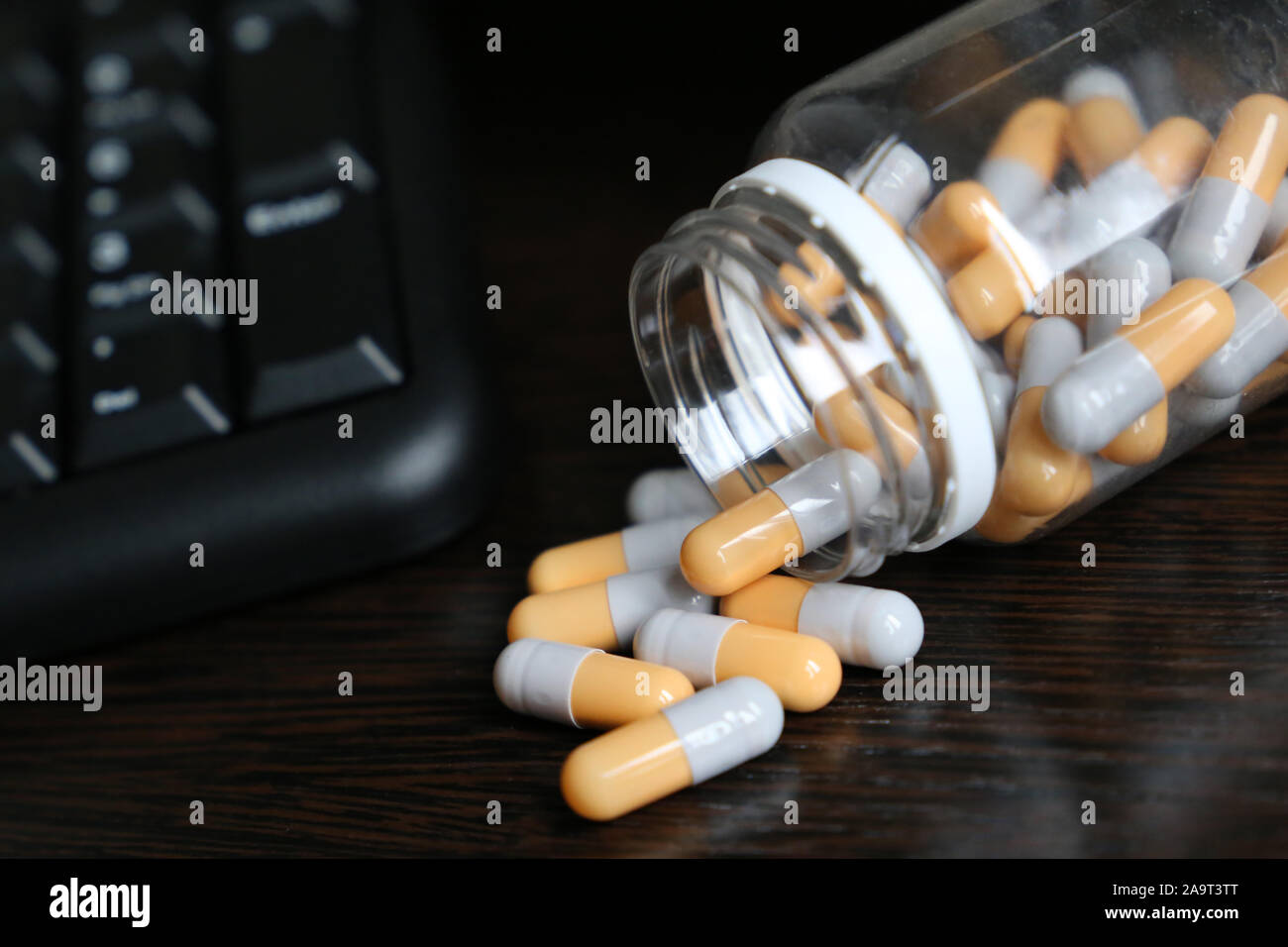 Pills near PC keyboard on a dark wooden table, capsules scattered from a bottle. Concept of taking medication in office, vitamins for energy and work Stock Photo