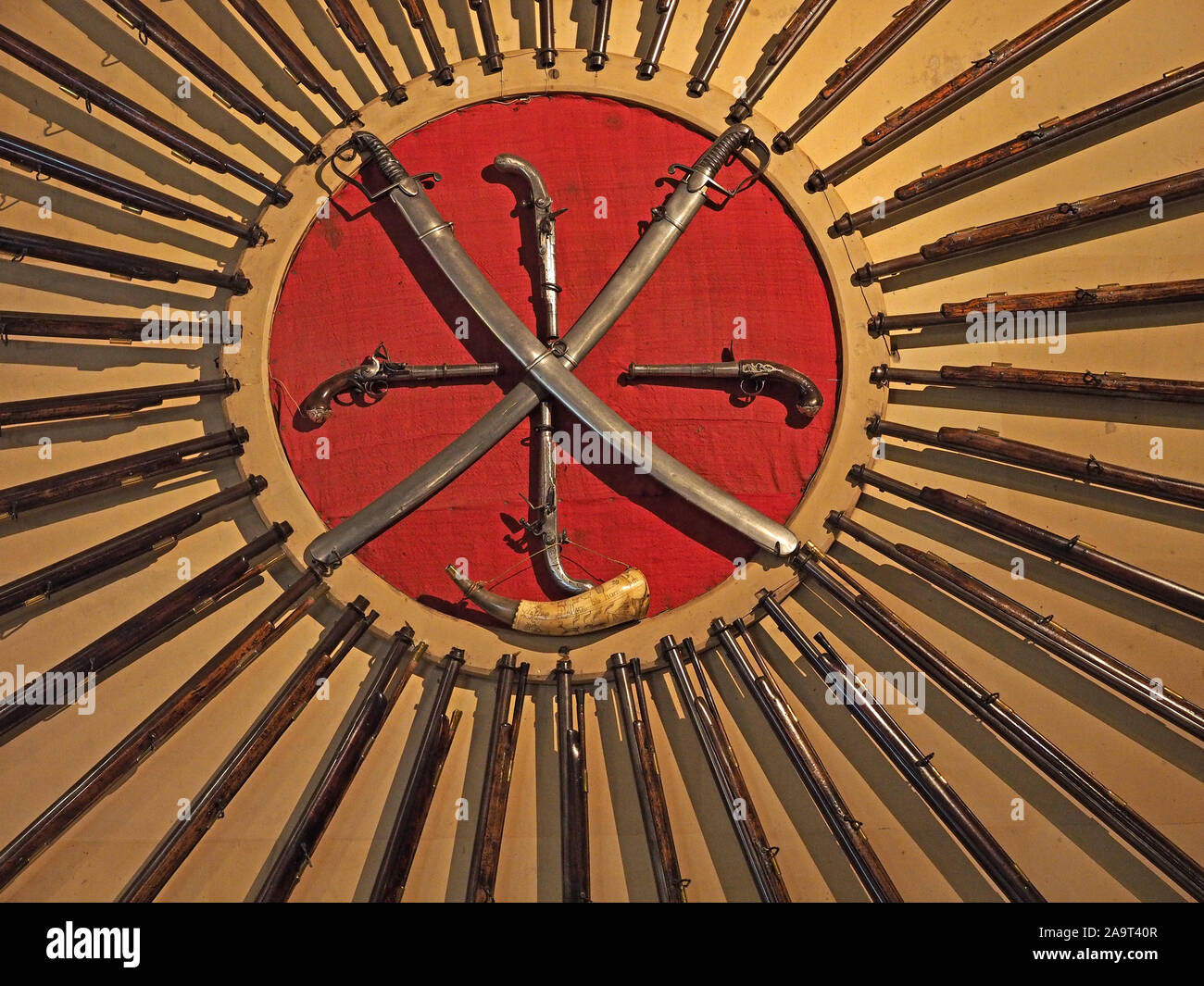 wheel of many rifle barrels surround a red circle with crossed sheathed swords & ancient pistols with powder horn create radial pattern  England, UK Stock Photo
