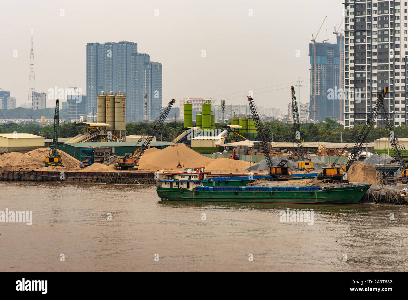 Ho Chi Minh City Vietnam - March 12, 2019: Song Sai Gon river. Green barge and many cranes and heaps of yellow sand on shore at ongoing base where con Stock Photo