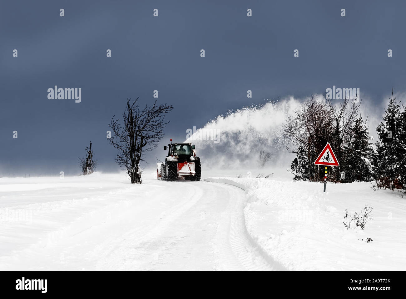 A tractor working as a snow plough uncovers a snow-covered road, the snow flies away in a high arc, dark sky, sign for cyclists - Location: border reg Stock Photo
