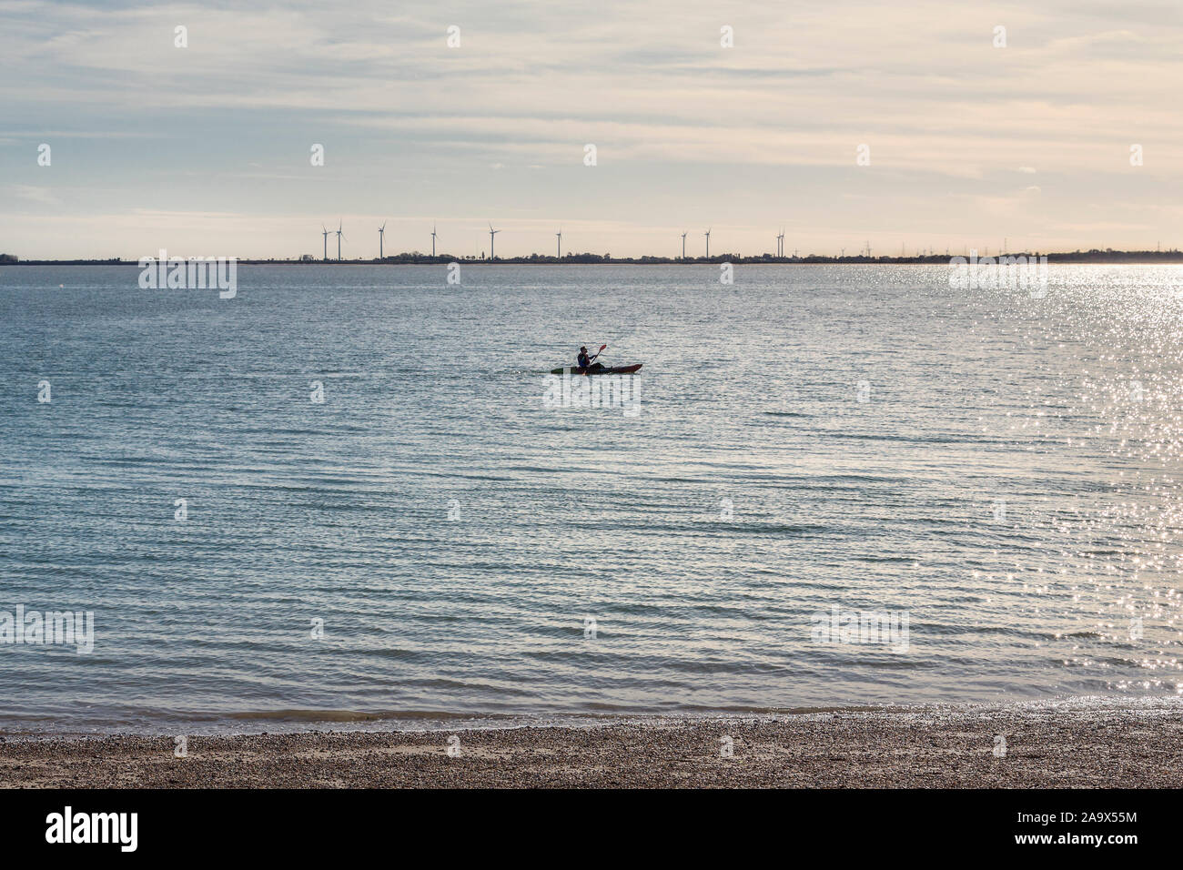 A kayaker paddles through the sea, behind him wind turbines can be seen. Possibly suggesting sustainable energy awareness and environmentalism. Stock Photo