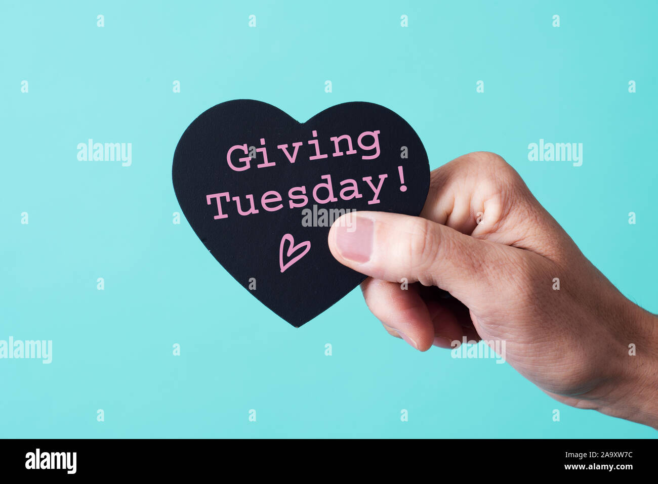 the hand of a young caucasian man holding a black heart-shaped sign, with the text giving tuesday written in it, on a pink background Stock Photo