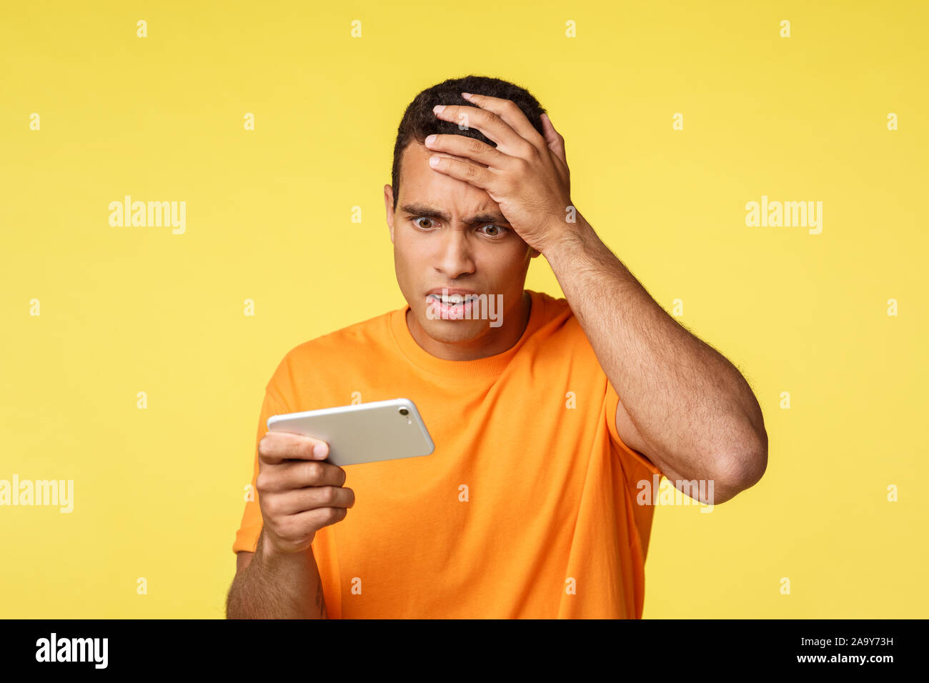 Bothered and upset shocked young handsome man, holding smartphone horizontally watch devastating news or video, punch forehead, frowning, lost in game Stock Photo