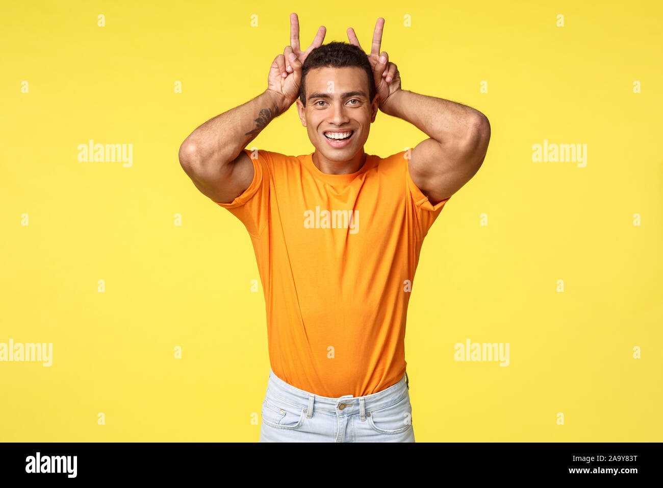 Cute masculine and silly hispanic boyfriend in orange t-shirt, make peace gesture, bunny ears behind head, smiling joyfully, mimick rabbit to fool Stock Photo