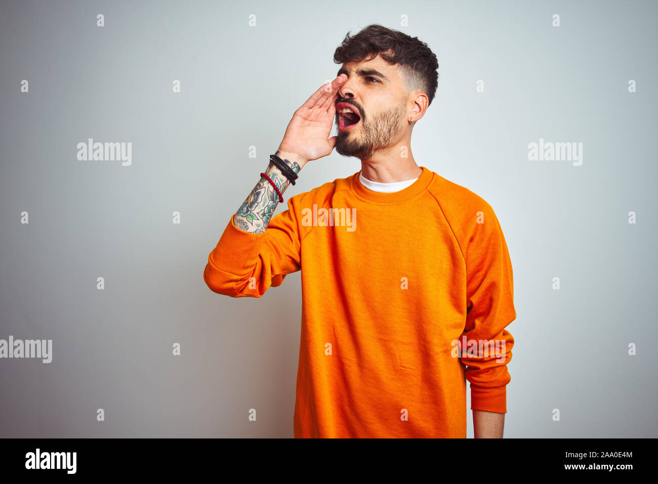 Young man with tattoo wearing orange sweater standing over isolated white background shouting and screaming loud to side with hand on mouth. Communica Stock Photo