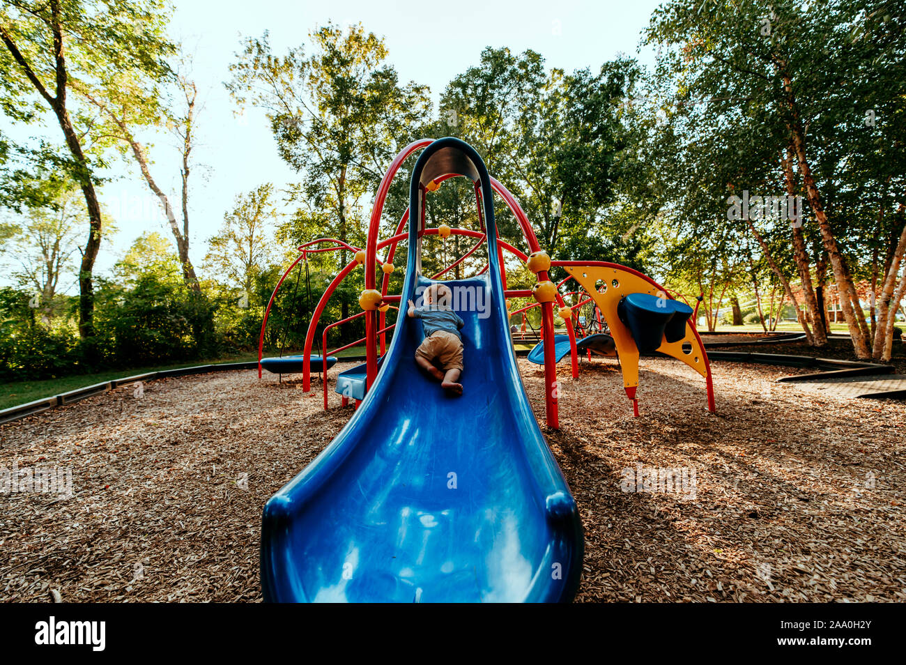 Baby boy climbing slide at a park with lots of trees and sunlight. Stock Photo