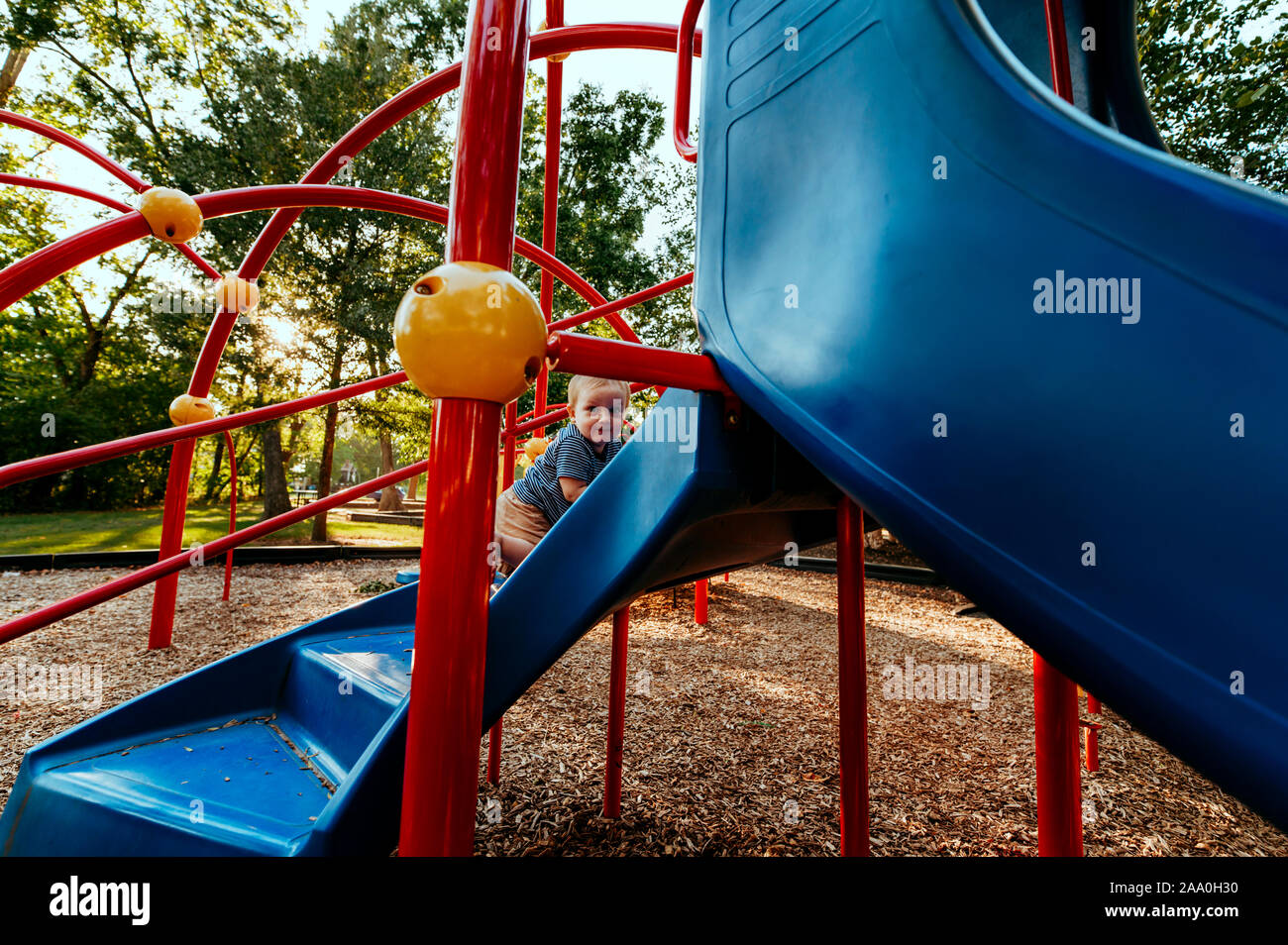 Toddler boy climbing stairs to blue slide at park with trees. Stock Photo