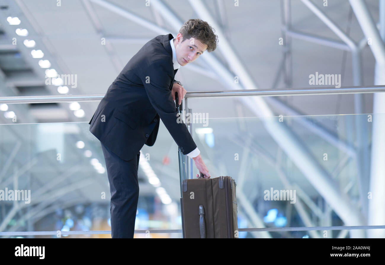 Young businessman wearing a suit standing at the glass guard railing inside an airport with his hands at his rolling suitcase Stock Photo