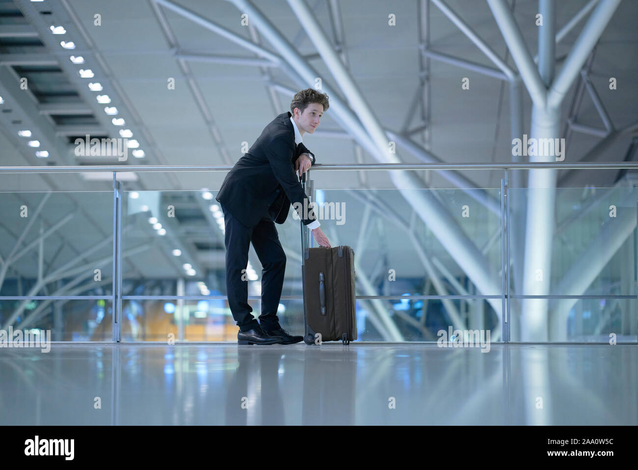 Young handsome businessman wearing a suit standing at the glass guard railing inside an airport with his hands at his rolling suitcase Stock Photo