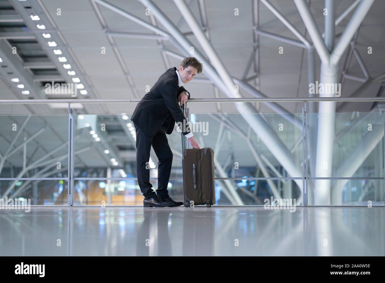 Young handsome businessman wearing a suit standing at the glass guard railing inside an airport with his hands at his rolling suitcase Stock Photo
