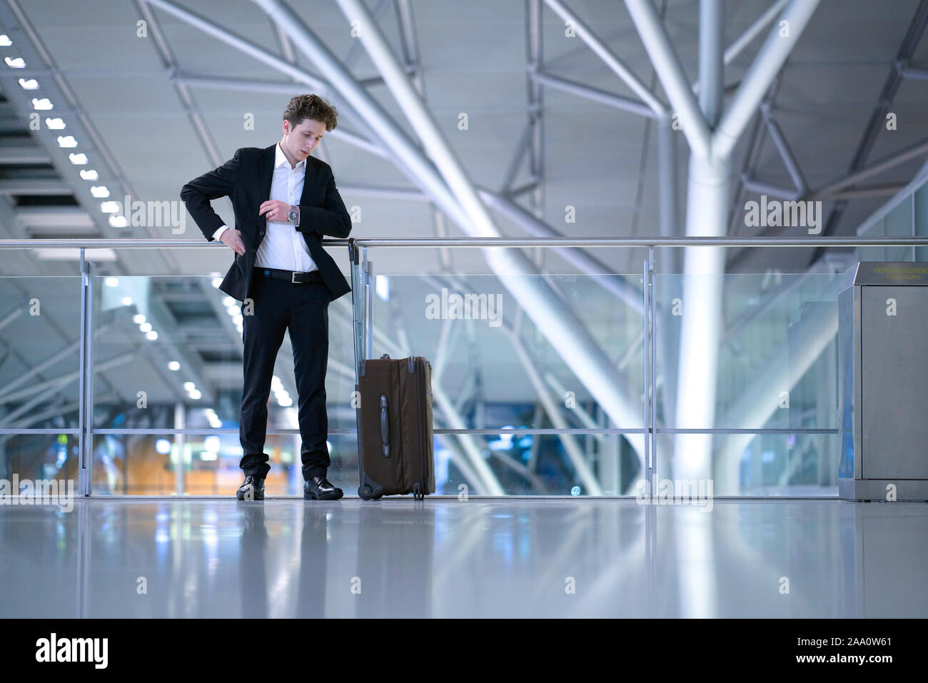 Young businessman standing inside the airport in front of a glass guard railing with his rolling case, searching something in his suit pocket Stock Photo