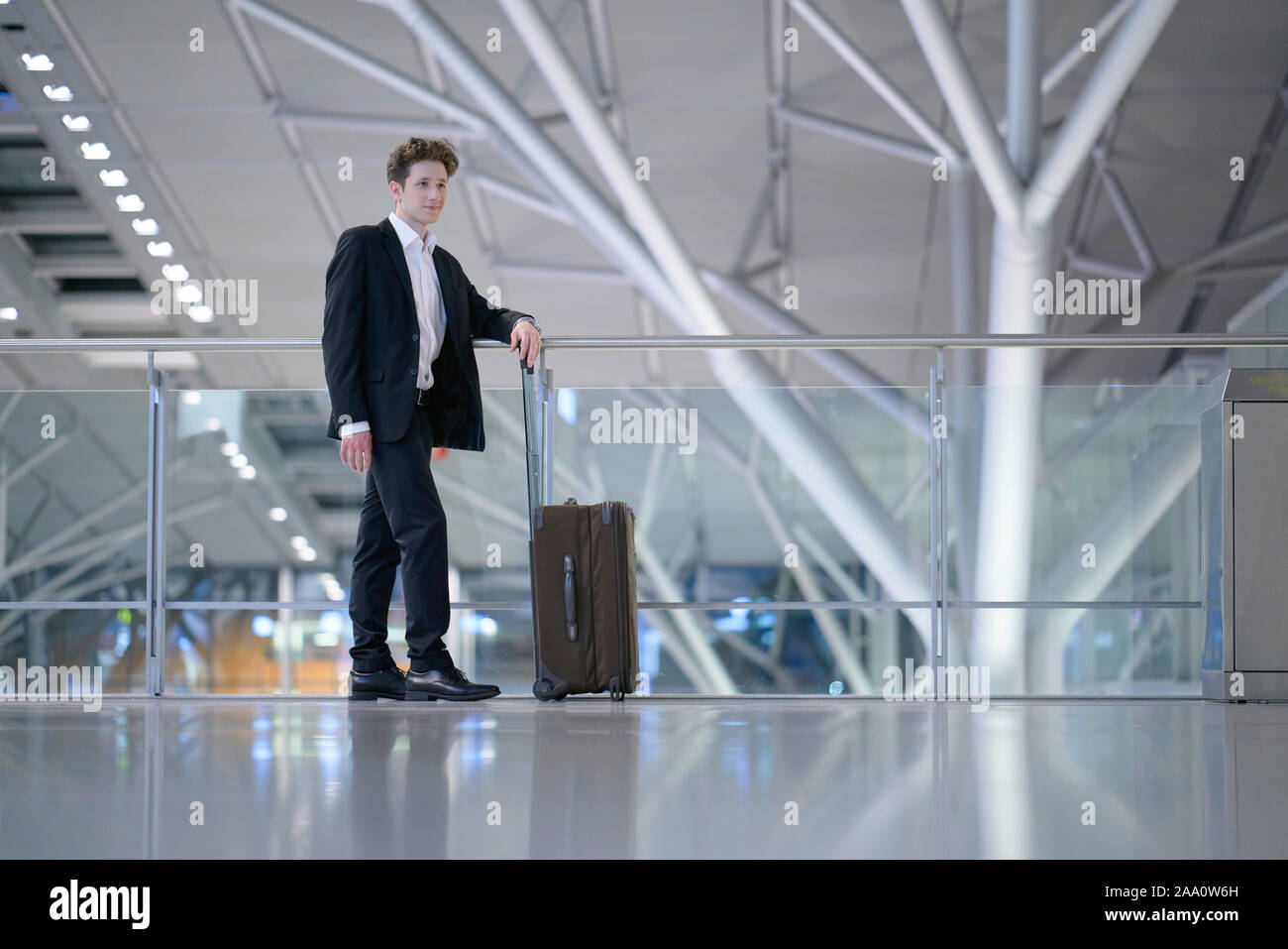 Young relaxed smiling businessman standing with his rolling suitcase in the airport, leaning with his arm on a guard railing Stock Photo