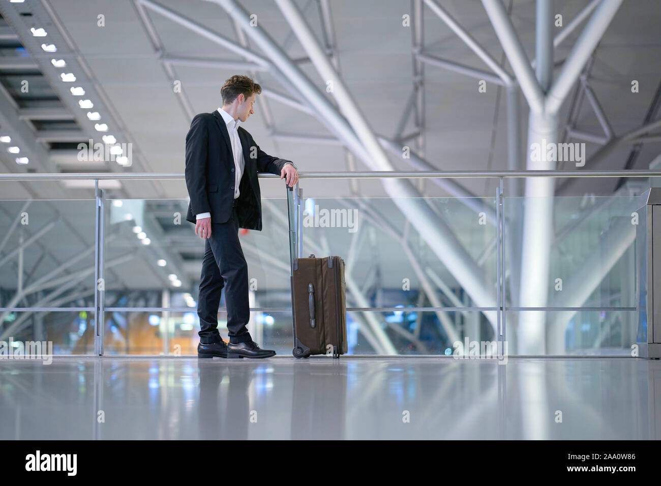 Young relaxed smiling businessman standing with his rolling suitcase in the airport, leaning with his arm on a guard railing and looking down Stock Photo