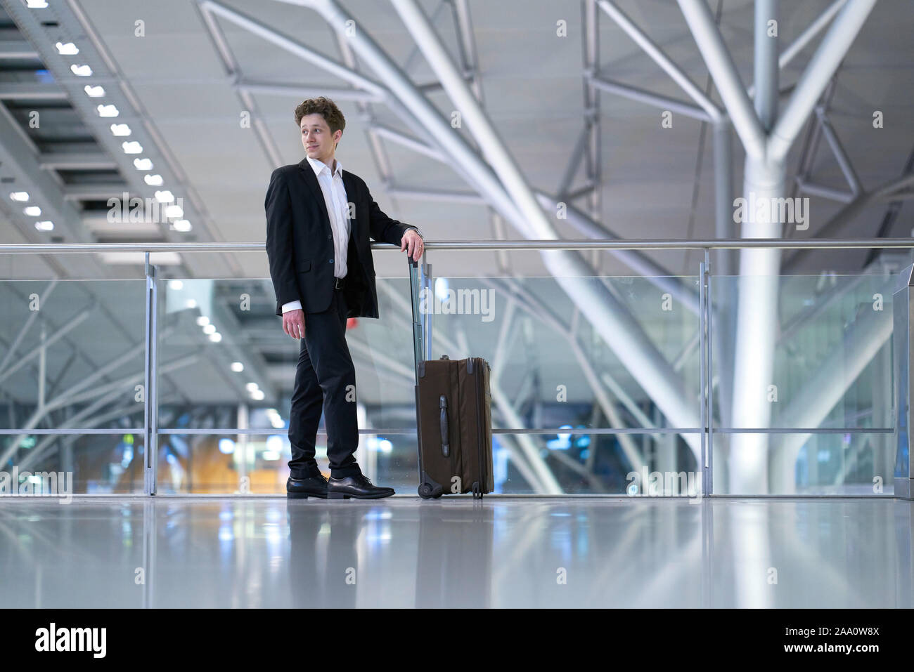 Young relaxed smiling businessman standing with his rolling suitcase in the airport, leaning with his arm on a guard railing, looking over his right s Stock Photo