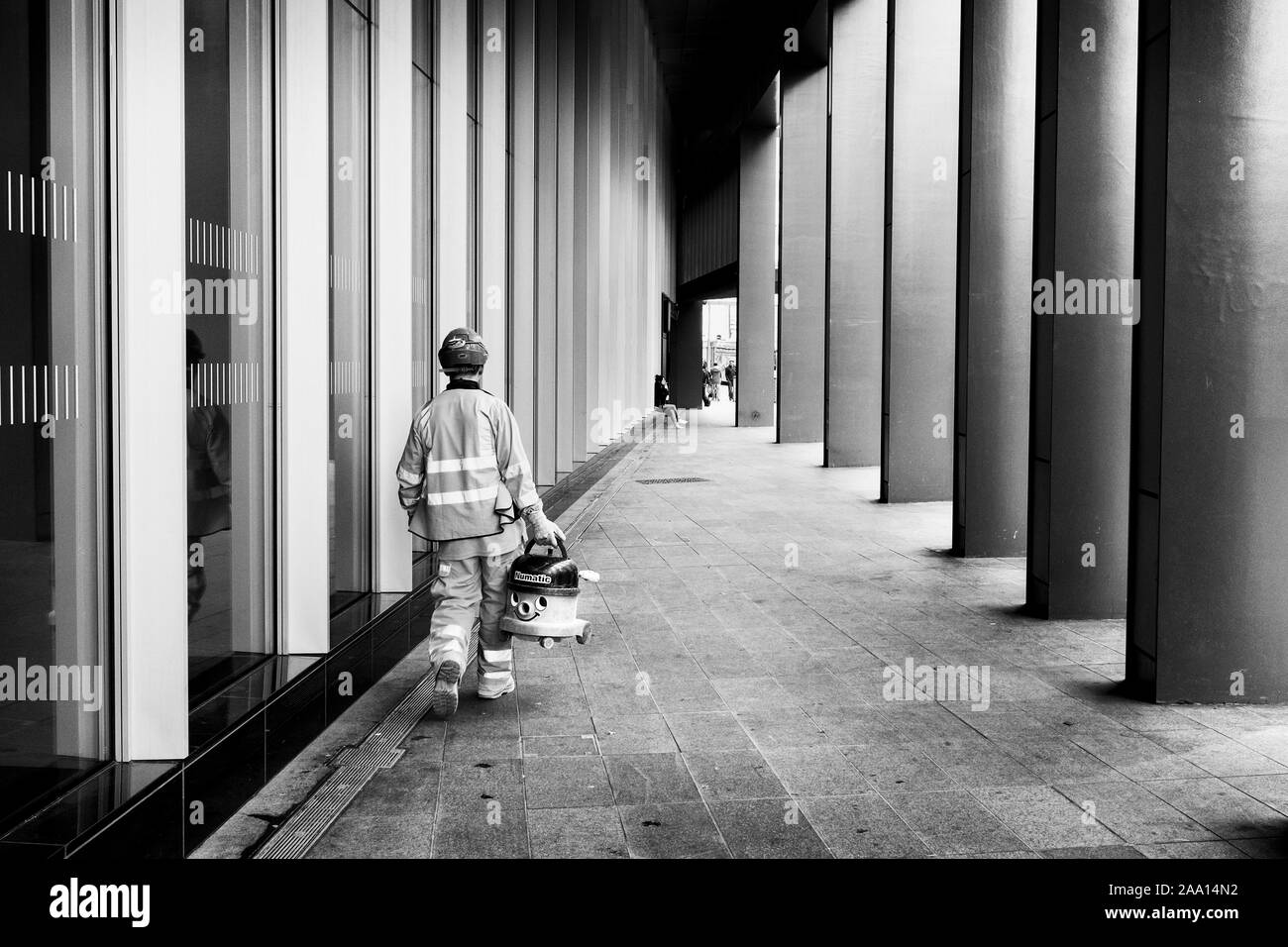 London black and white street photography: Workman carrying Numatic 'Henry' vacuum cleaner in street. Stock Photo