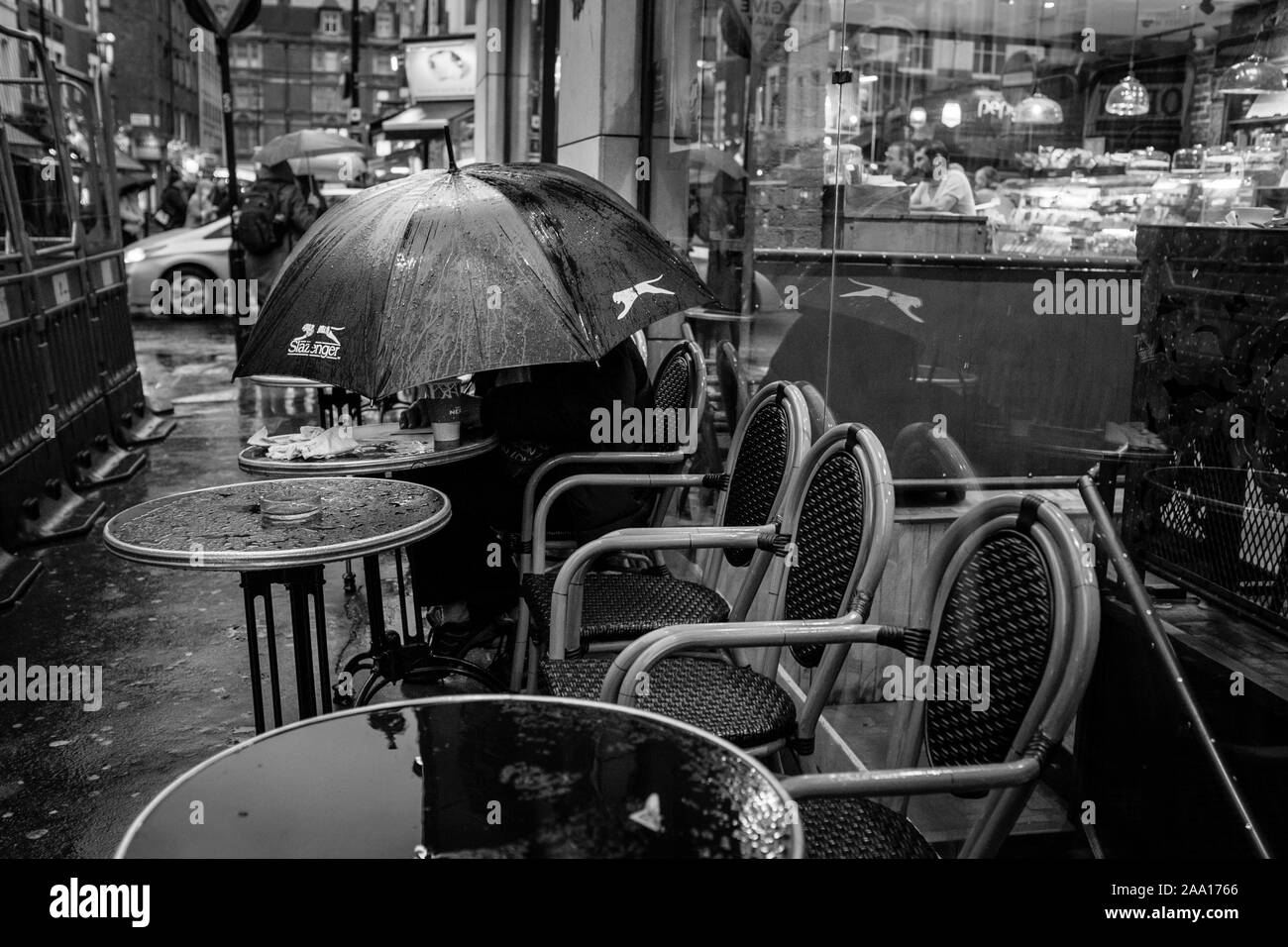 London black and white street photography: Customer sits outside cafe under umbrella during downpour, Frith street, London, UK Stock Photo