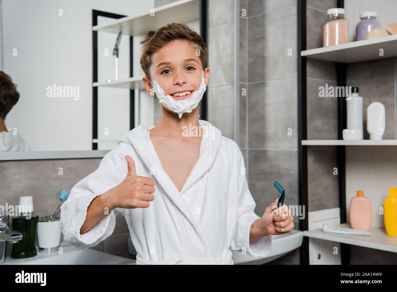 happy boy with shaving foam on face holding razor and showing thumb up ...