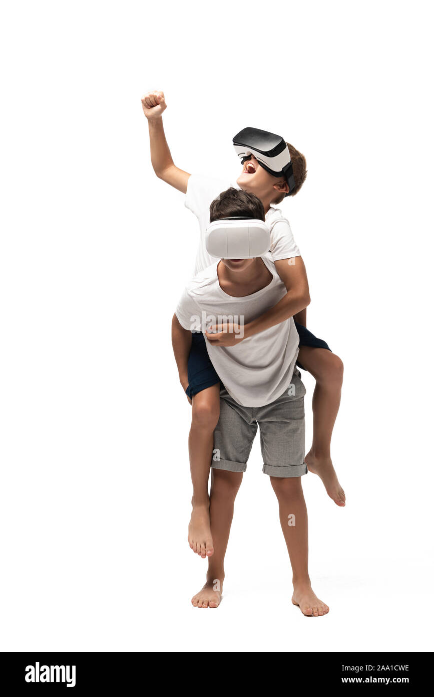 cheerful boy piggybacking on brothers back and showing yes gesture while using vr headsets together on white background Stock Photo