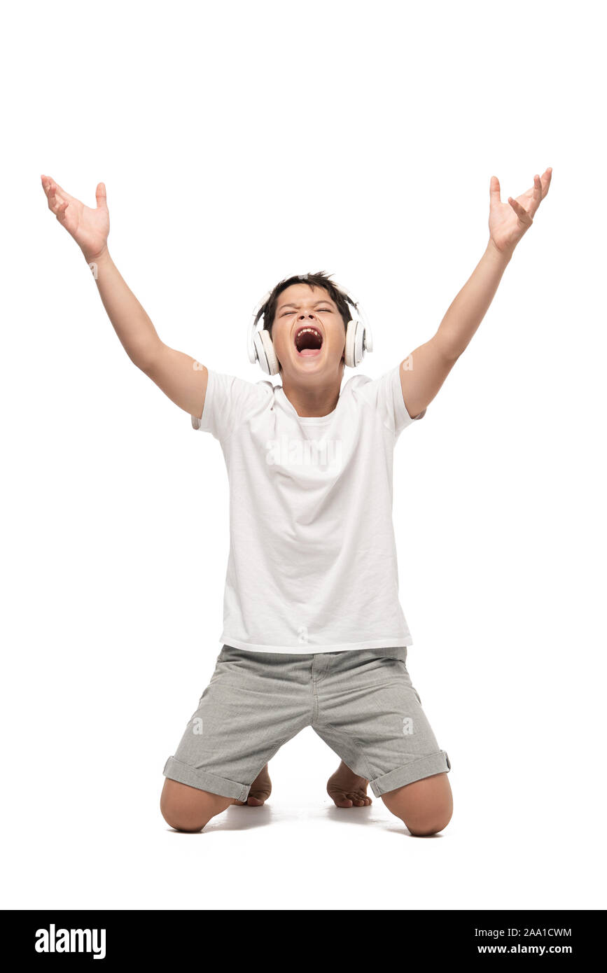 excited boy standing on knees with raised hands while listening music in headphones and singing on white background Stock Photo