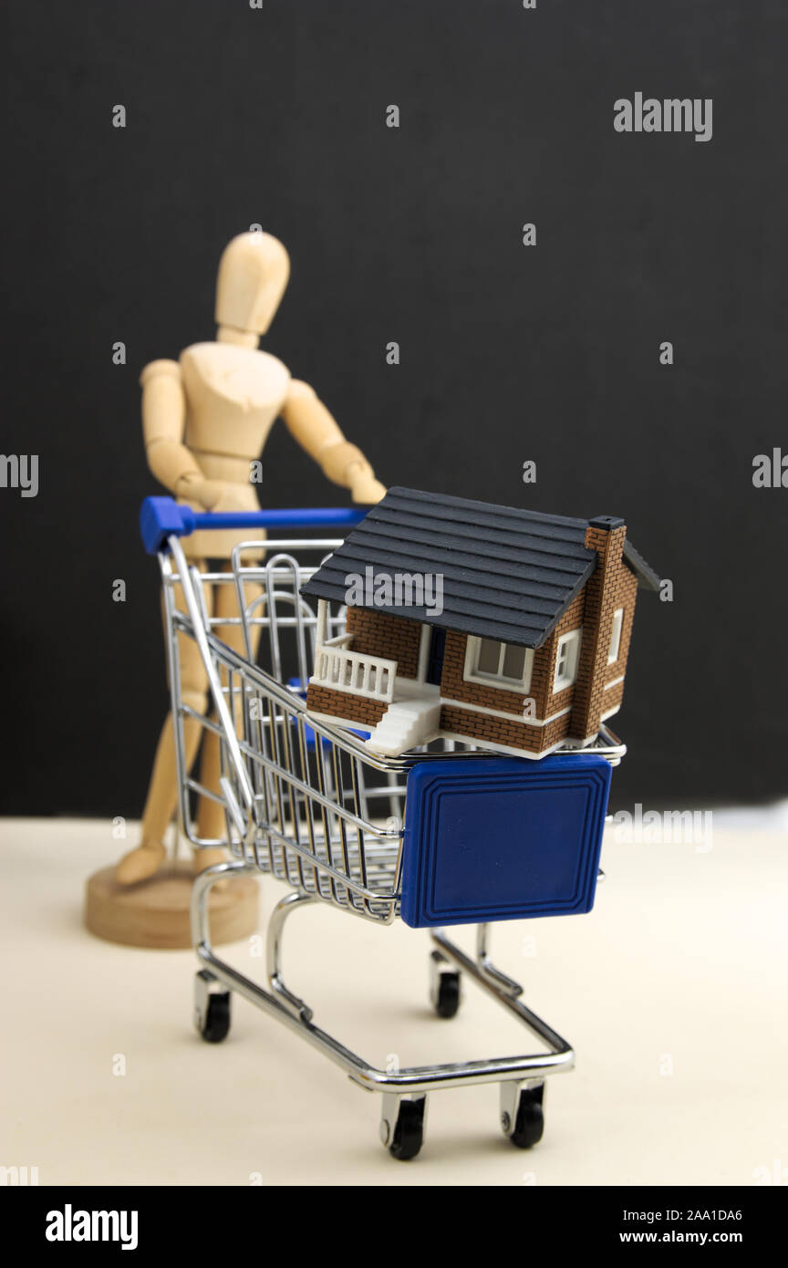 A dummy carries a shopping cart with a miniature house with a dark background. Concept of buying houses. Stock Photo