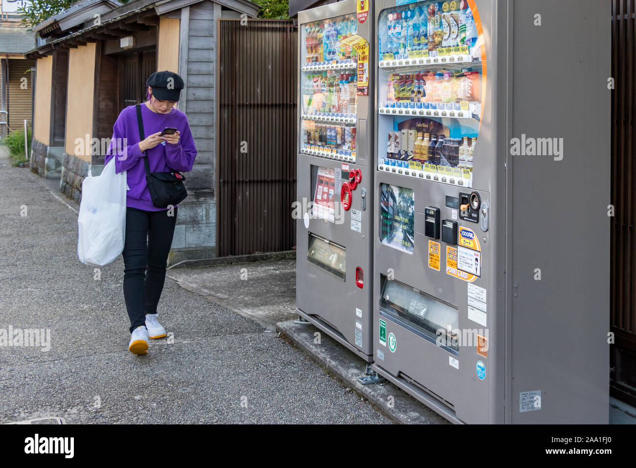 A young japanese woman texting - chating with her mobile next to a automatic drinks vendor machine in Kanazawa, Japan. Stock Photo
