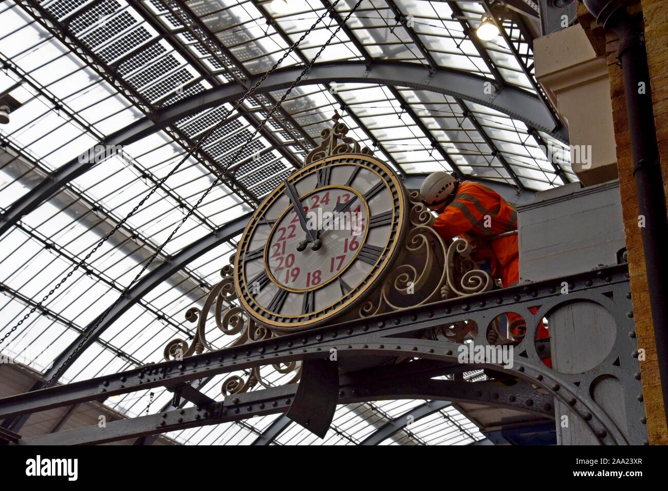 A clock repairer attends to maintenance on the large station clock above the platform at London Kings Cross Station - Stock Image