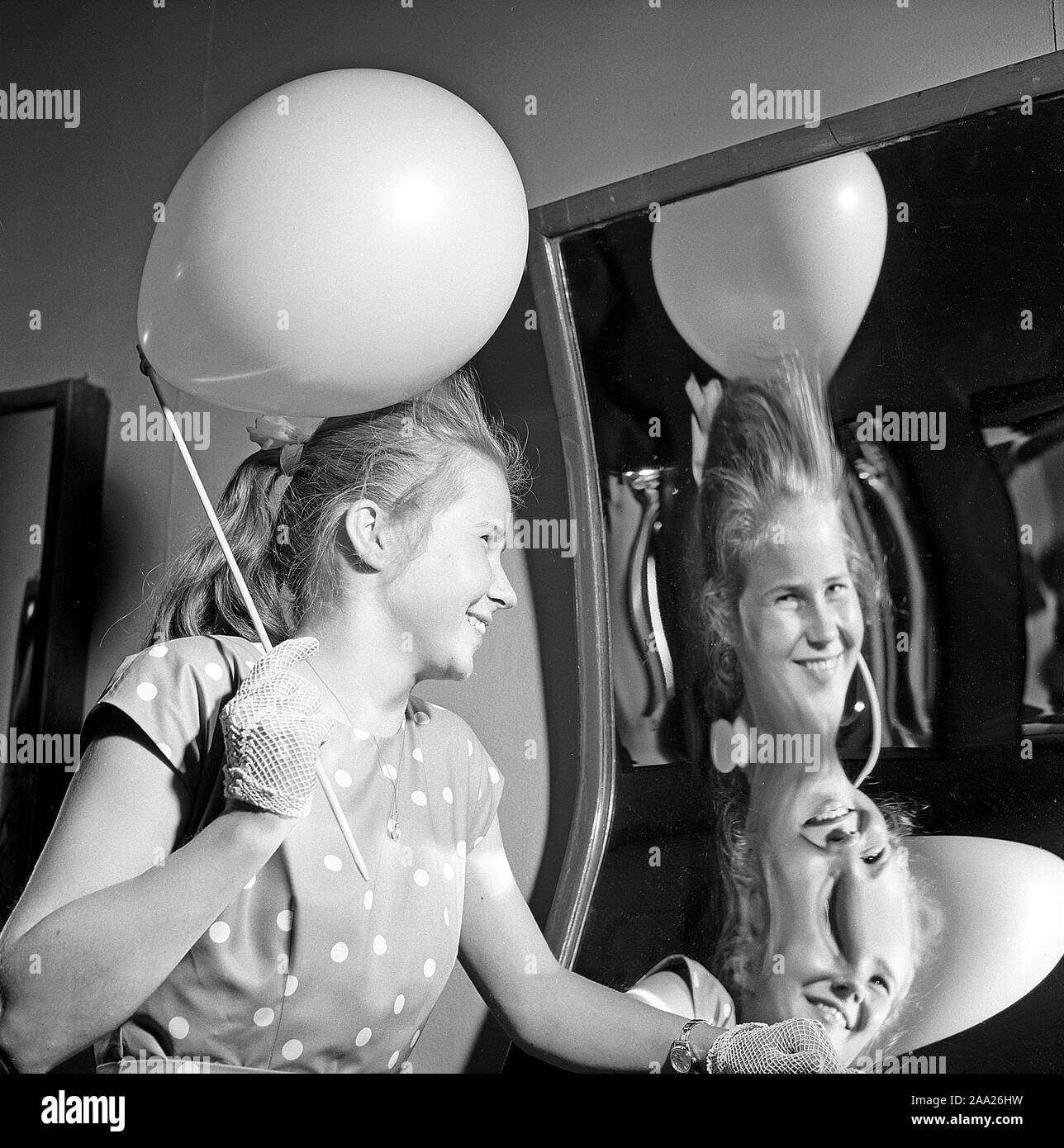 Amusement park in the 1950s. A teenage girl is laughing at how she look in  a funny mirror. Mirrors with different shapes makes the reflection of  people look tall and thin, or