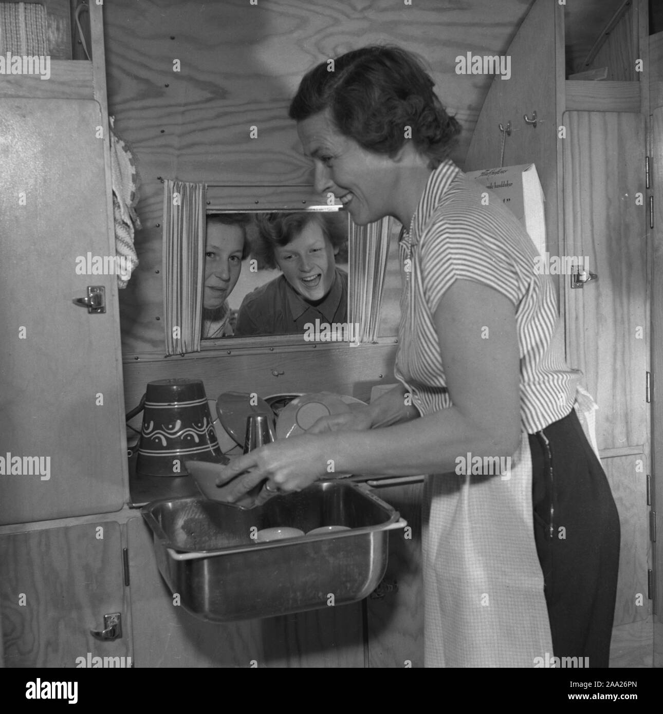 1950s camping. A family is enjoying their holiday and the practial camping life in their caravan. Demonstrating how well everything works for them even on holiday. The mother is doing the dishes in the practical small kitchen sink, with her children cheering on from outside the window. Sweden 1952 Ref 2021 Stock Photo