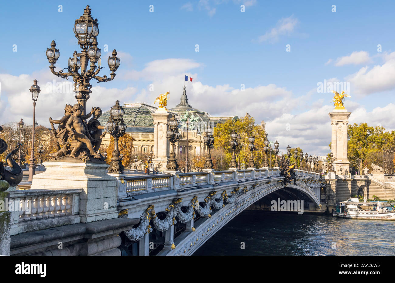 Famous Pont Alexandre III bridge on sunny autumn day. Behind is roof of Grand Palais with French flag waiving against clouds Stock Photo