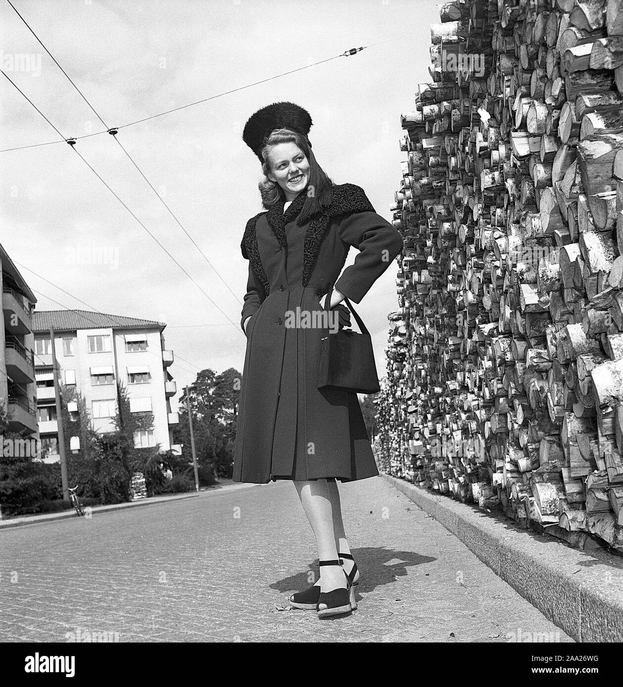 Women's fashion in the 1940s. A young woman in a typical 1940s coat with matching hat, shoes and handbag. It's postwar Sweden and there is still a shortage of fuel and coal for heating, and the piles of firewood is visible in the background. Kristoffersson Ref V78-6. Sweden 1947 Stock Photo