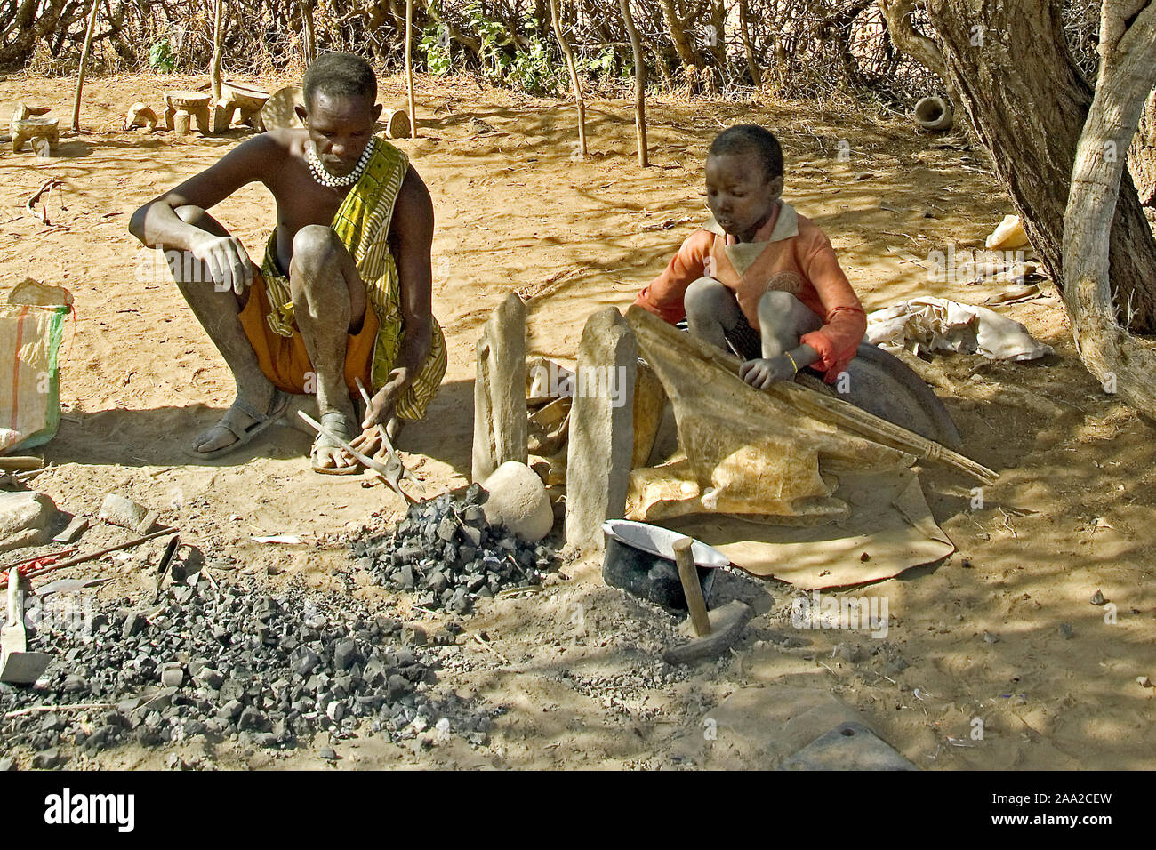 Local blacksmith of Datoga Tribe and his children working in the shade of an Acasia tree.  Lake Eyasi, northern Tanzania. Stock Photo