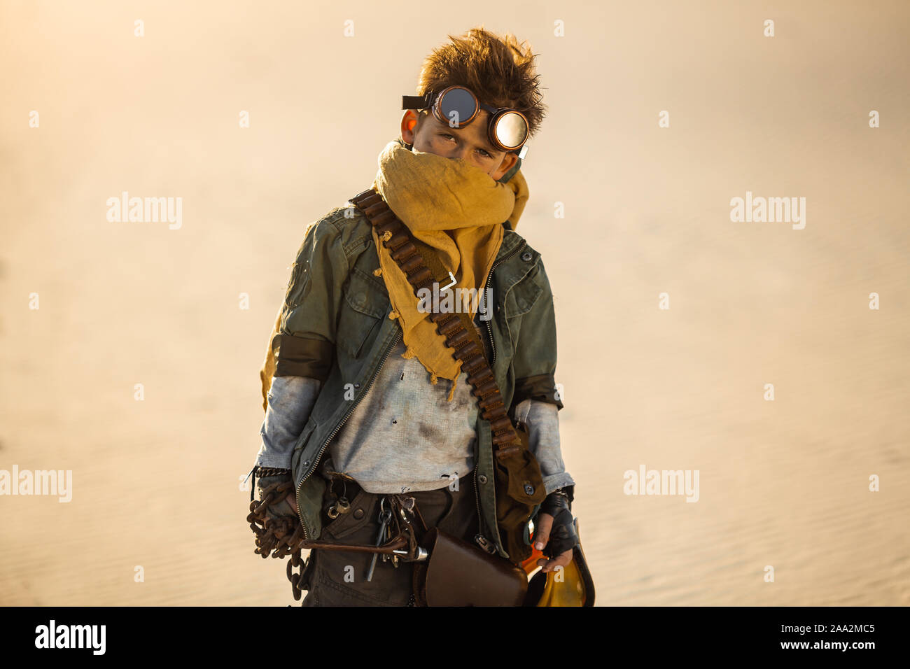 Post-apocalyptic cyberpunk boy outdoors. Nuclear post-apocalypse time. Life  after doomsday concept. A young man warrior with chain, gas mask, in shabby  clothes standing in an aggressive pose, desert and dead wasteland on