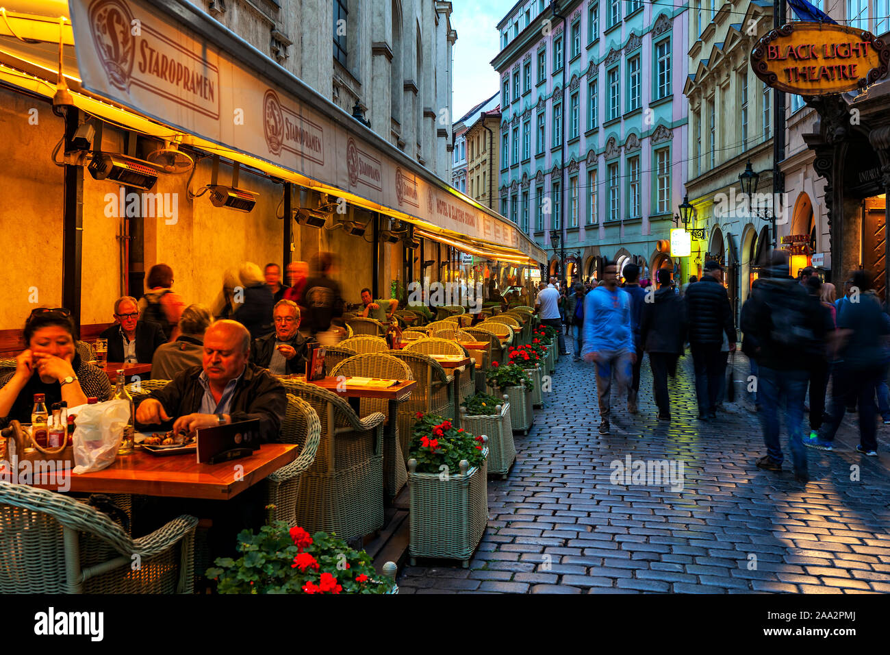 People sitting in illuminated outdoor restaurant next to crowd cobblestone street in Old Town of Prague. Stock Photo