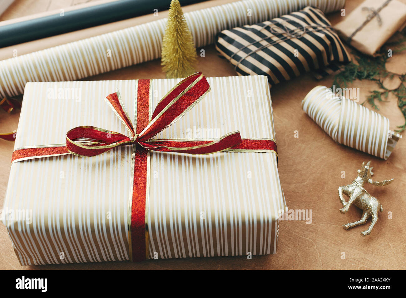 https://c8.alamy.com/comp/2AA2XKY/stylish-christmas-gift-box-in-striped-golden-paper-and-with-red-bow-and-scissors-presents-golden-tree-deer-on-wooden-table-wrapping-christmas-gif-2AA2XKY.jpg