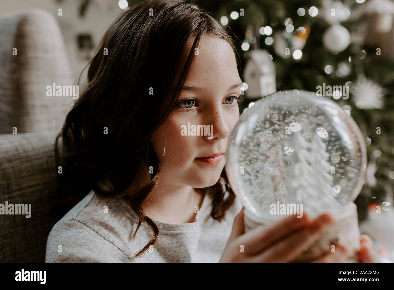 Girl sitting by a Christmas tree looking at a snow globe Stock Photo