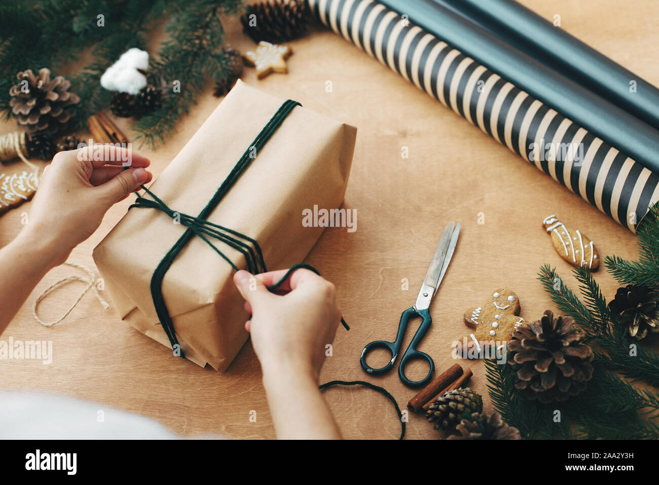 Hands wrapping rustic christmas gift, tying green twine ,and pine branches, cones, gingerbread cookies, thread, cinnamon on rural wooden table. Wrappi Stock Photo