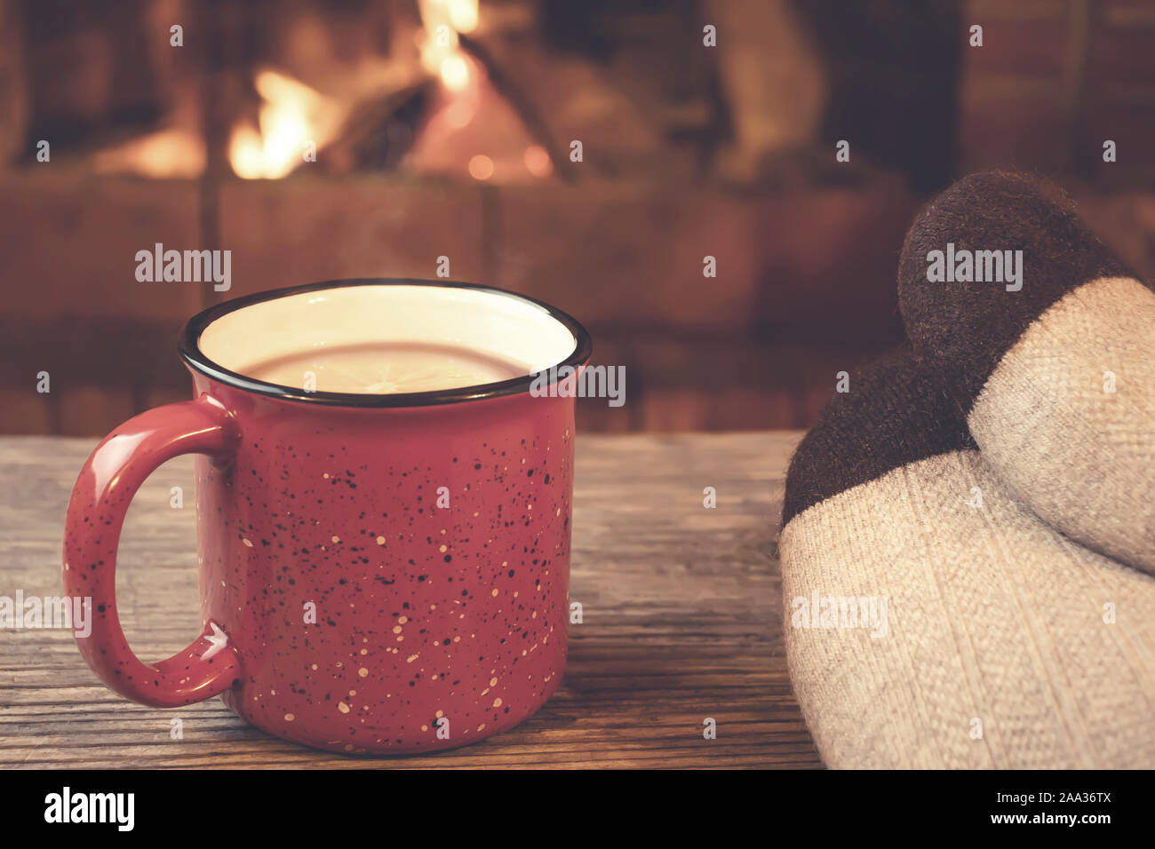 Feet in woolen socks, a red mug with hot tea opposite the burning fireplace, comfort, and warmth of the hearth concept. Stock Photo