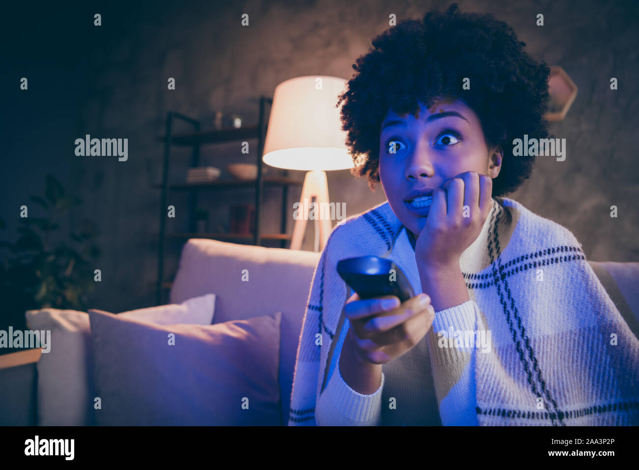 Close up photo of terrified afro american girl sit divan watch horror film hold remote control switch channel feel nervous bite nail cover plaid Stock Photo