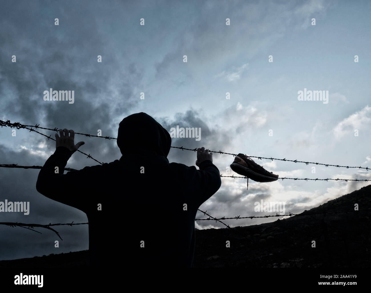 Rear view of man wearing hoodie behind barbed wire, razor wire fence. Shoe hanging from fence Stock Photo